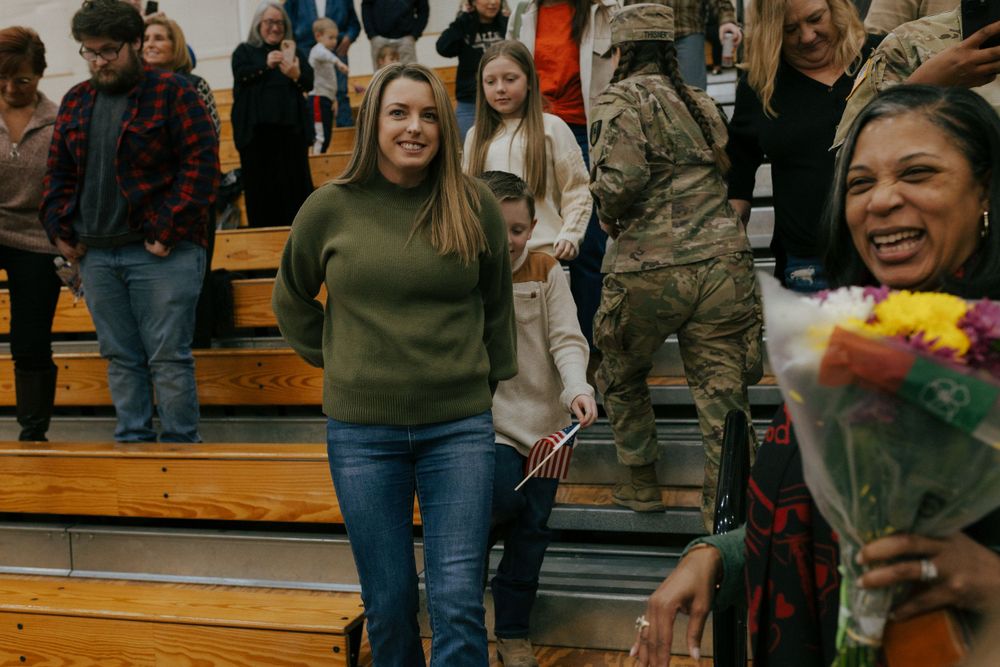 Woman in olive green turtleneck standing in crowded bleachers at an indoor event.