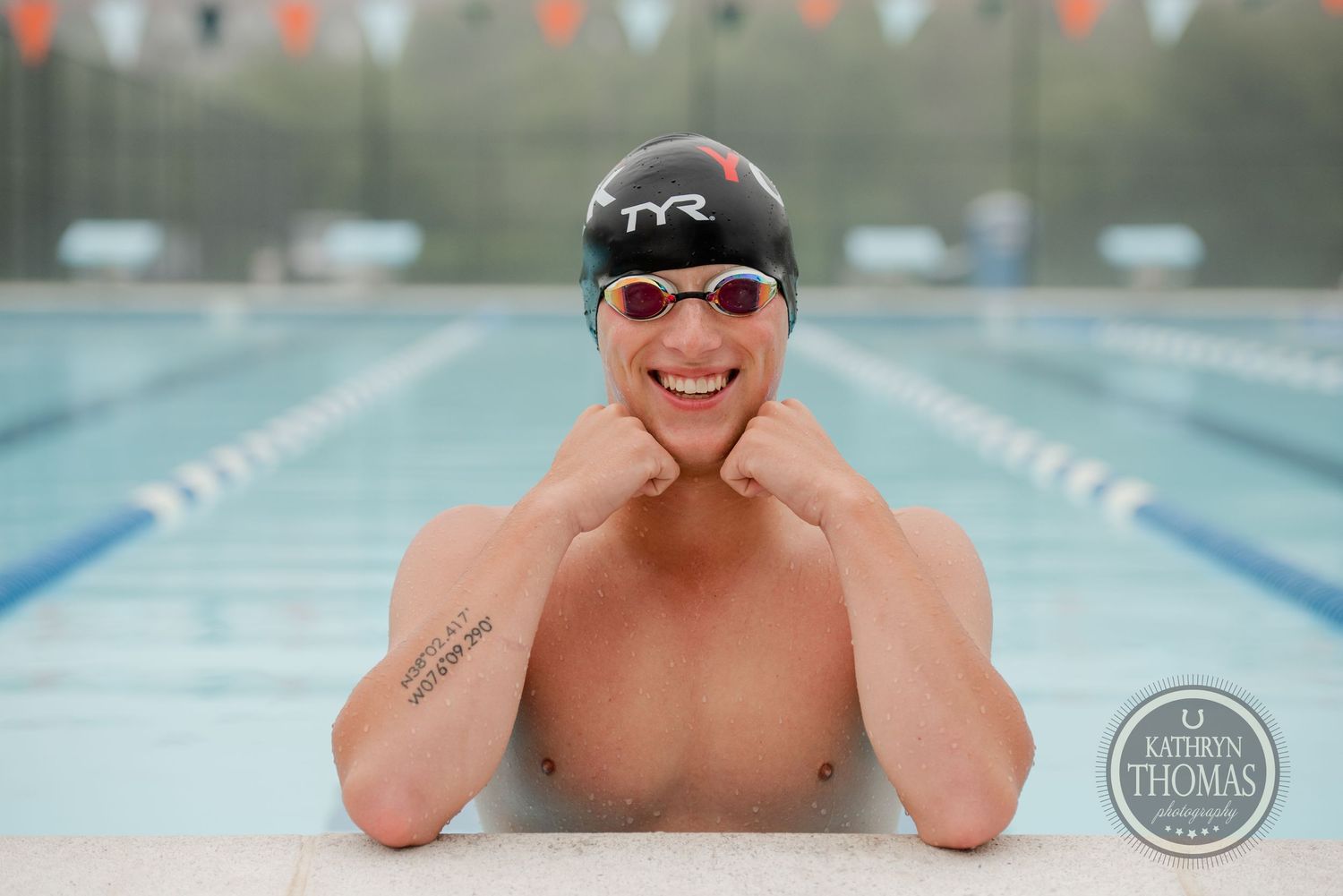 Cheerful swimmer in TYR cap and goggles posing at pool edge.