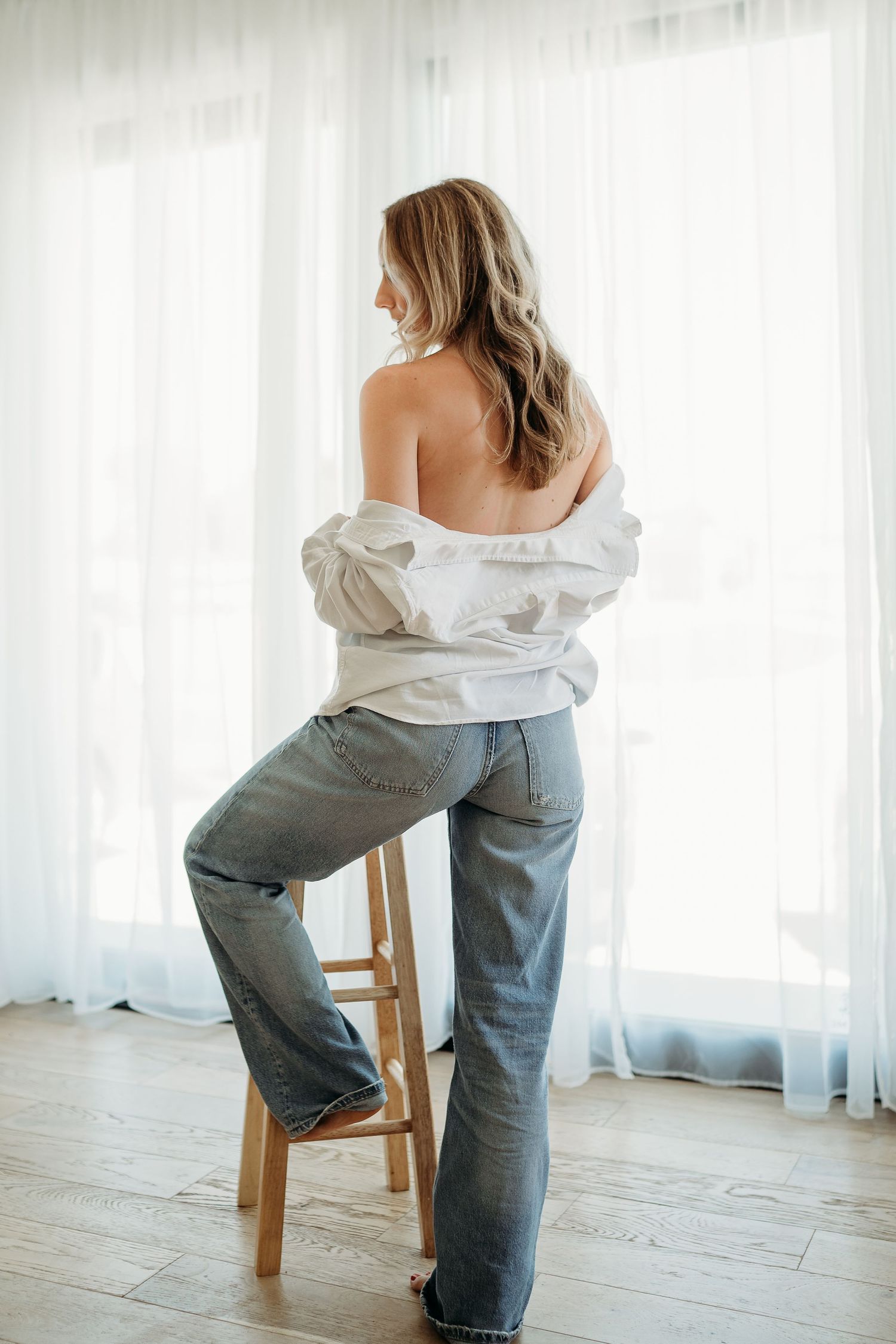 Artistic portrait of person in white shirt and jeans sitting on wooden stool against bright window.