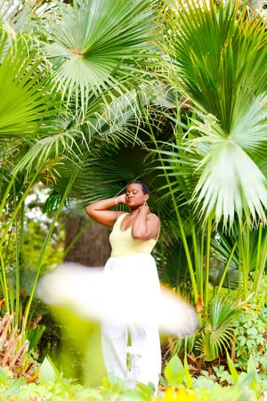 A woman in a yellow top and white pants poses gracefully among lush tropical fan palm trees in an outdoor garden setting.