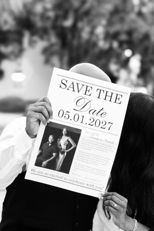 Black and white photo of a couple kissing behind a save the date card for a wedding on 05.01.2027 featuring their engagement portrait.