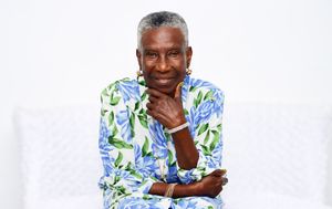 Elderly Black woman in floral blue-green shirt sits gracefully, smiling with hand on chin against white background.