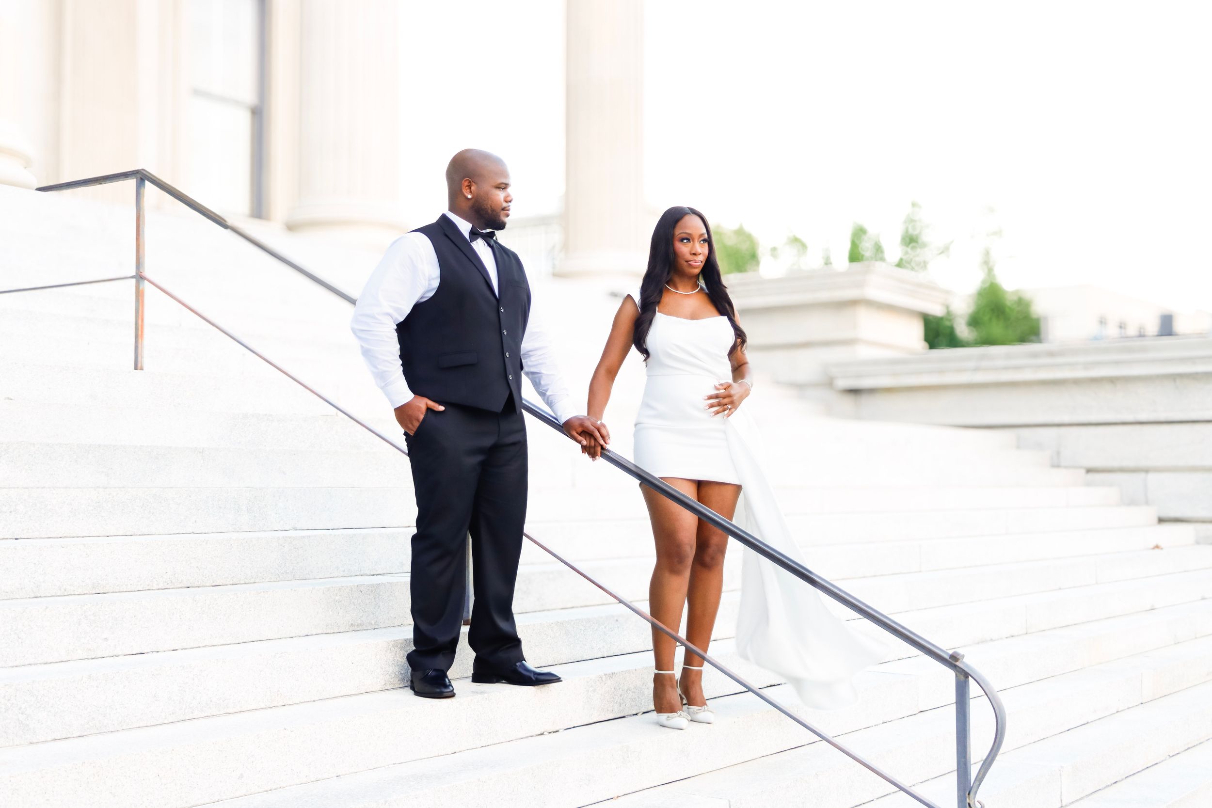 Couple in elegant attire pose on wide stone steps beside a metal railing, the man in a vest and bow tie, the woman in a white dress.