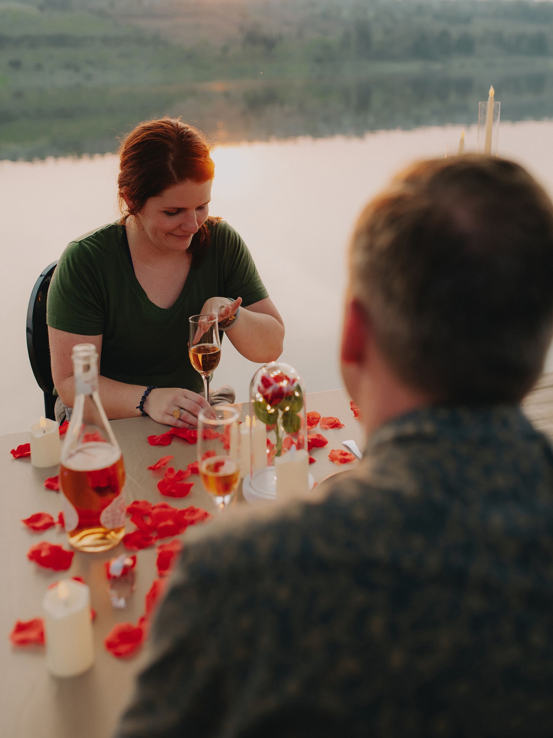 A romantic dinner setup with rose petals and drinks on a table by a serene lake at sunset.