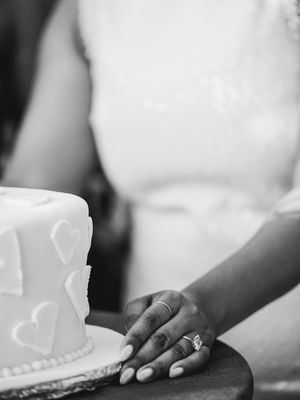Wedding cake with heart designs and a hand wearing an engagement ring resting beside it.