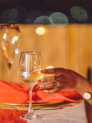 Hand wearing engagement ring holds wine glass against bokeh background.