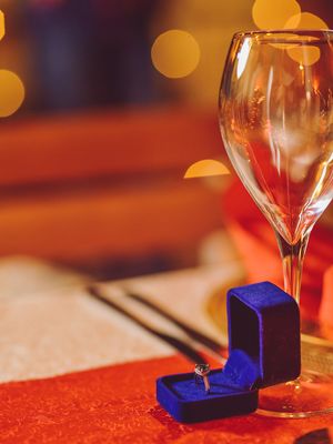 Engagement ring in blue velvet box next to wine glass on red tablecloth.