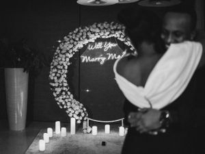 Silhouetted couple by a floral moon arch with 'Will You Marry Me' neon sign.