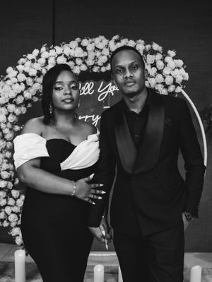 Elegant couple posing under floral arch with proposal sign in formal attire.