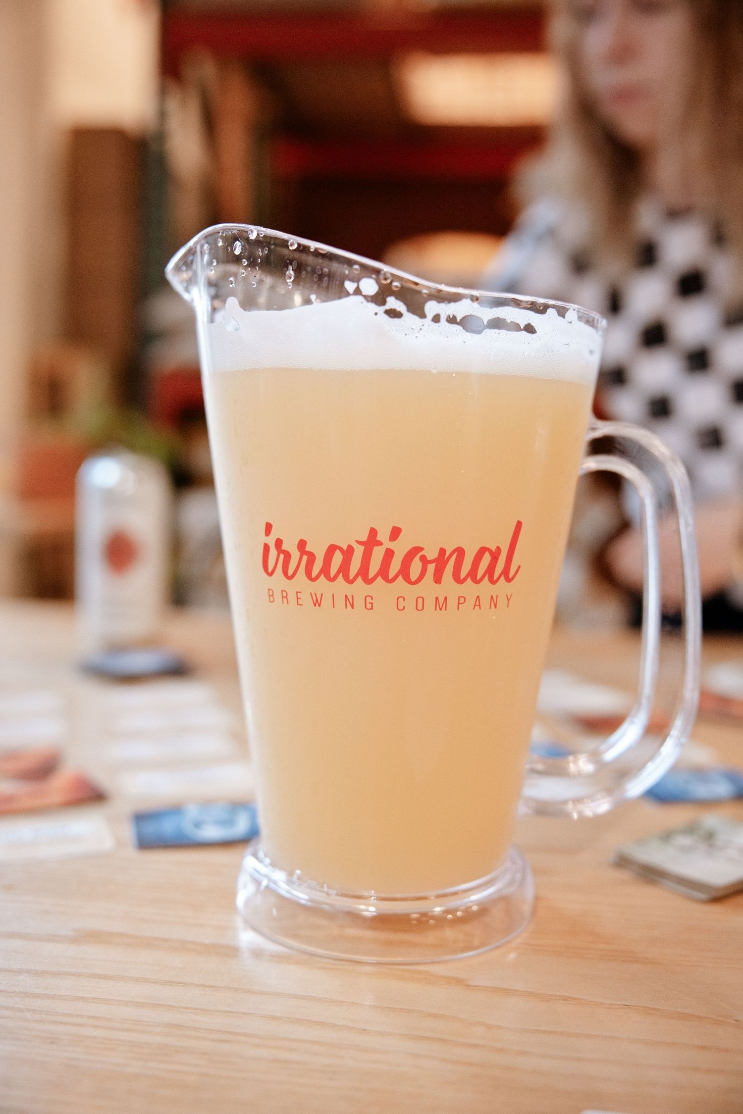 A glass pitcher of pale beer from Irrational Brewing Company sits on a wooden table in a casual setting.