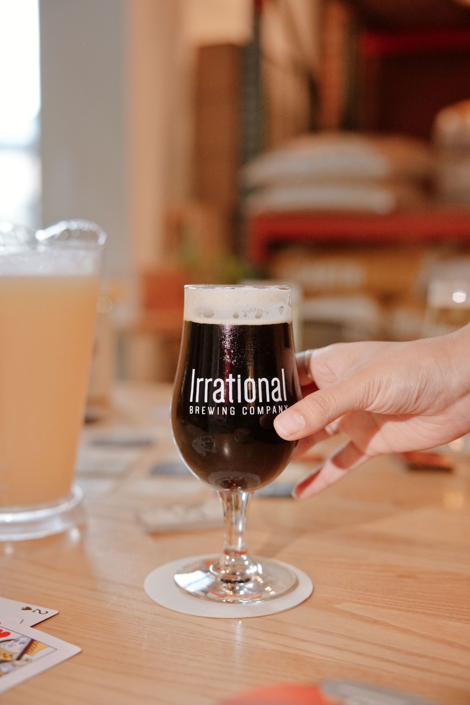 Hand holding a dark beer glass from Irrational Brewing Company on a wooden table in a taproom.