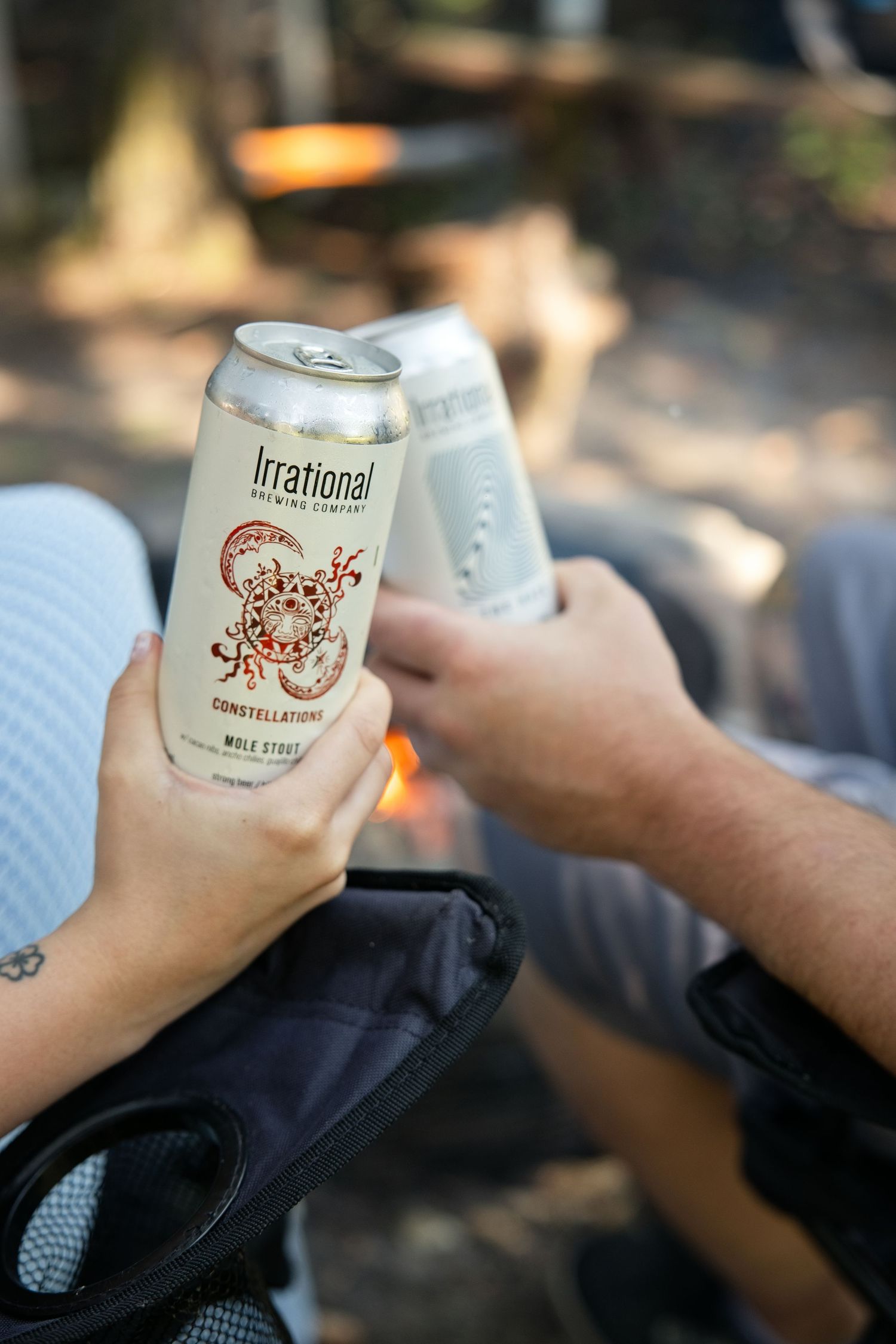 Two people clinking Irrational Brewing Company Constellations Milk Stout cans outdoors near a campfire.