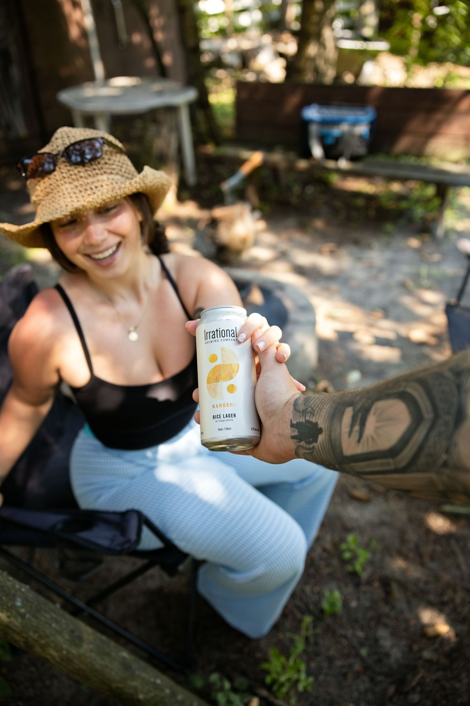 Woman in straw hat laughing outdoors, handing a can of Irrational beer to a tattooed hand.