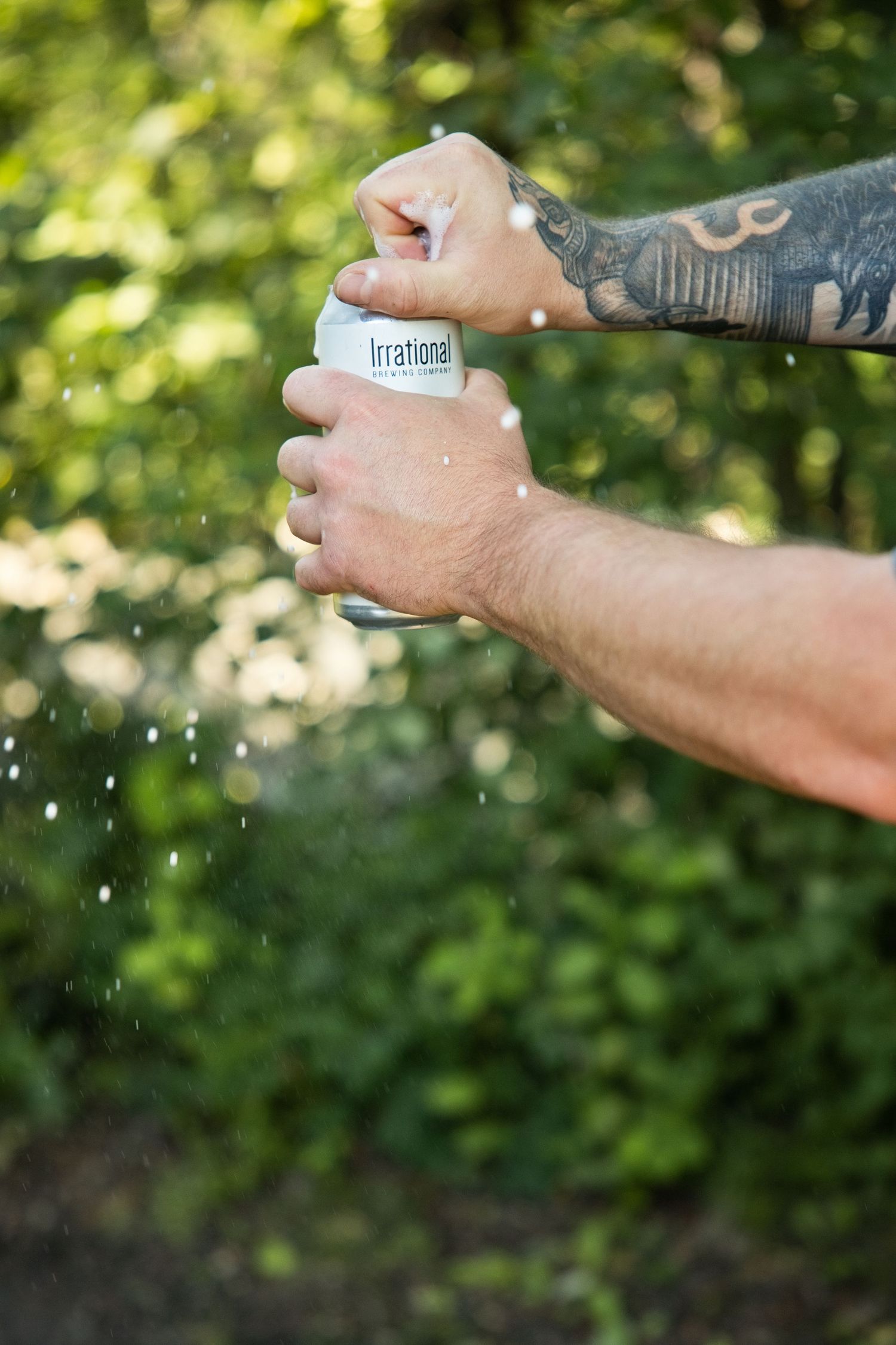 Tattooed hands opening a can labeled Irrational outdoors, with liquid spraying against a green leafy background.