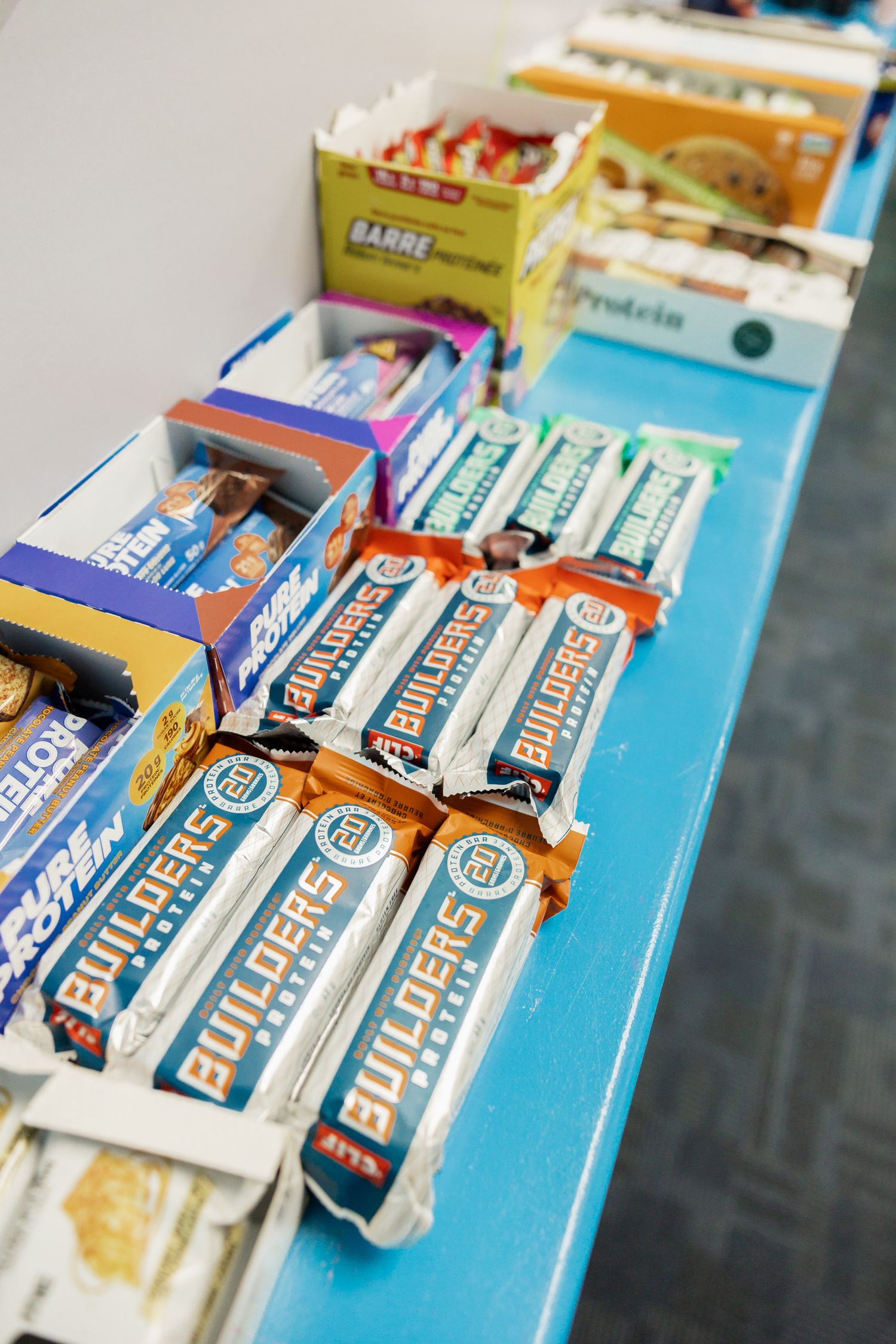 Assorted protein bars including Builders and Pure Protein displayed on a blue table at a fitness or nutrition event.