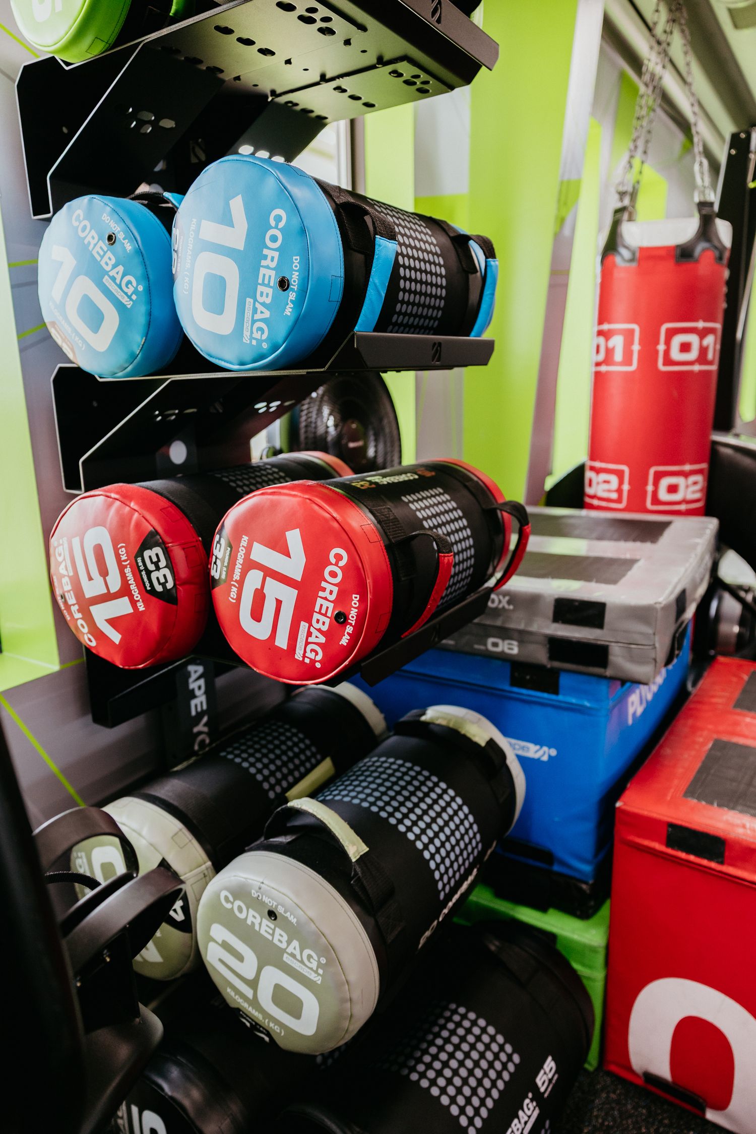 Rack of CoreBag weighted training bags in 10, 15, and 20kg with red punching bags in a gym.