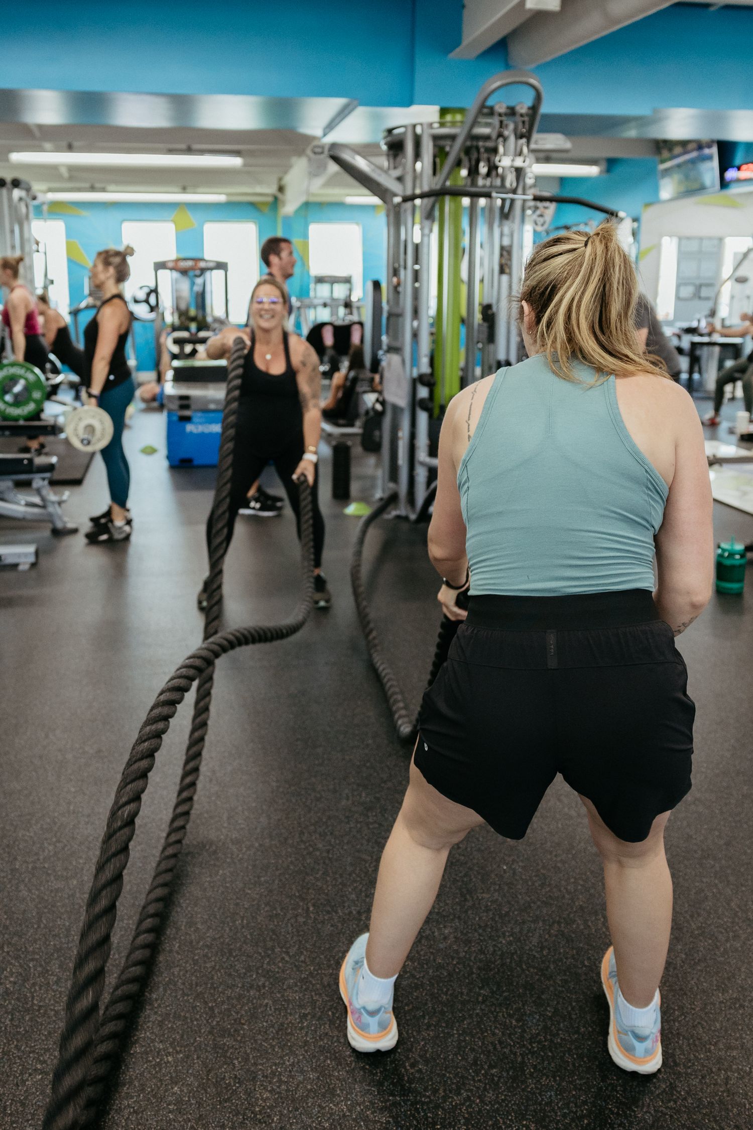 Two women doing battle rope exercises in a modern gym with blue walls and fitness equipment in the background.