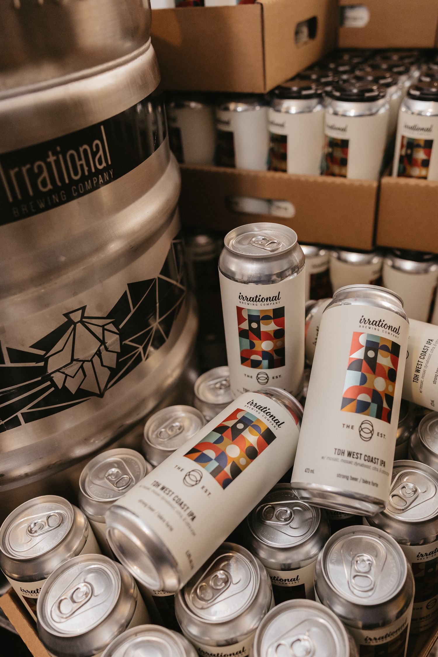 Irrational Brewing Company kegs and colorful geometric patterned beer cans stacked in a brewery storage area.