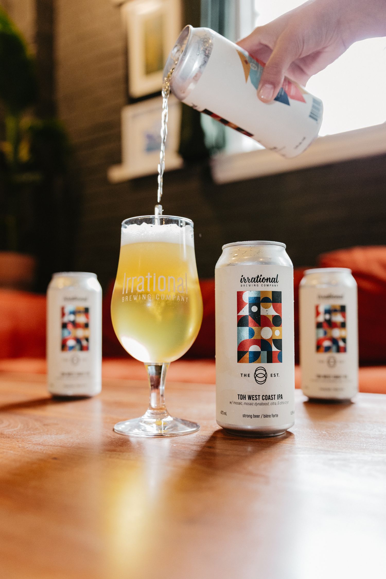 Hand pouring Irrational Brewing West Coast IPA can into a tulip glass on a wooden table with two cans nearby.