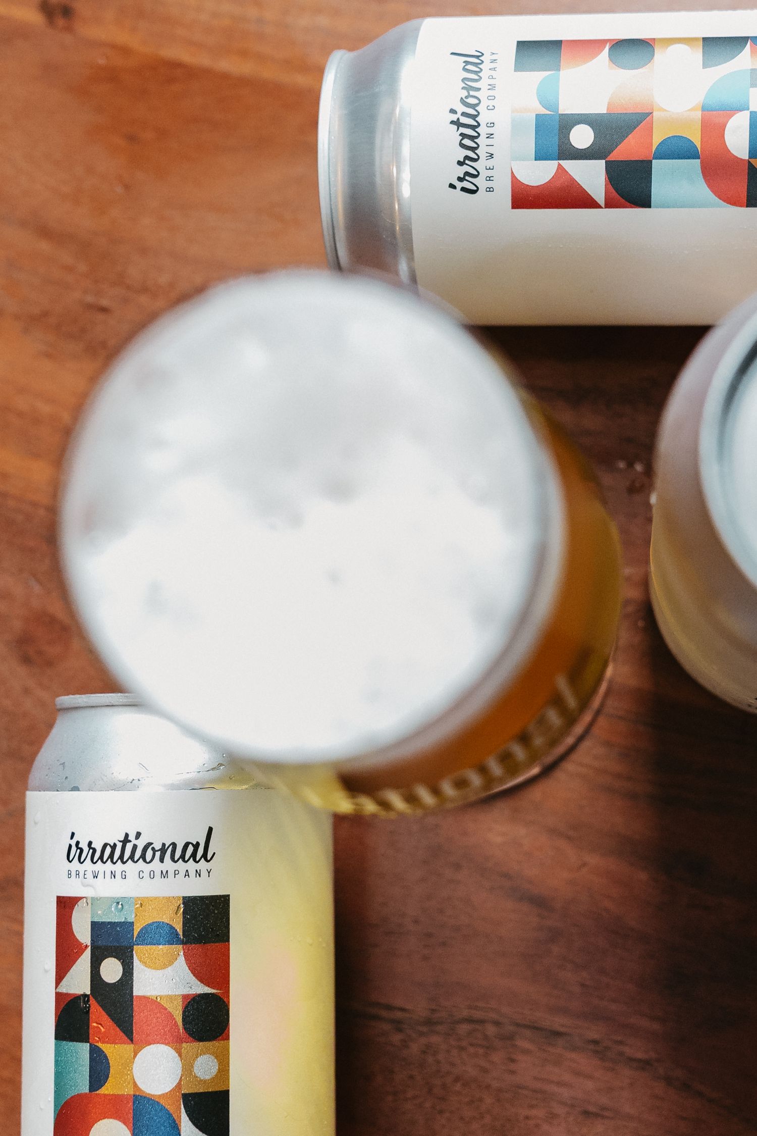 Irrational Brewing Company beer cans beside a frothy pint glass on a wooden table.