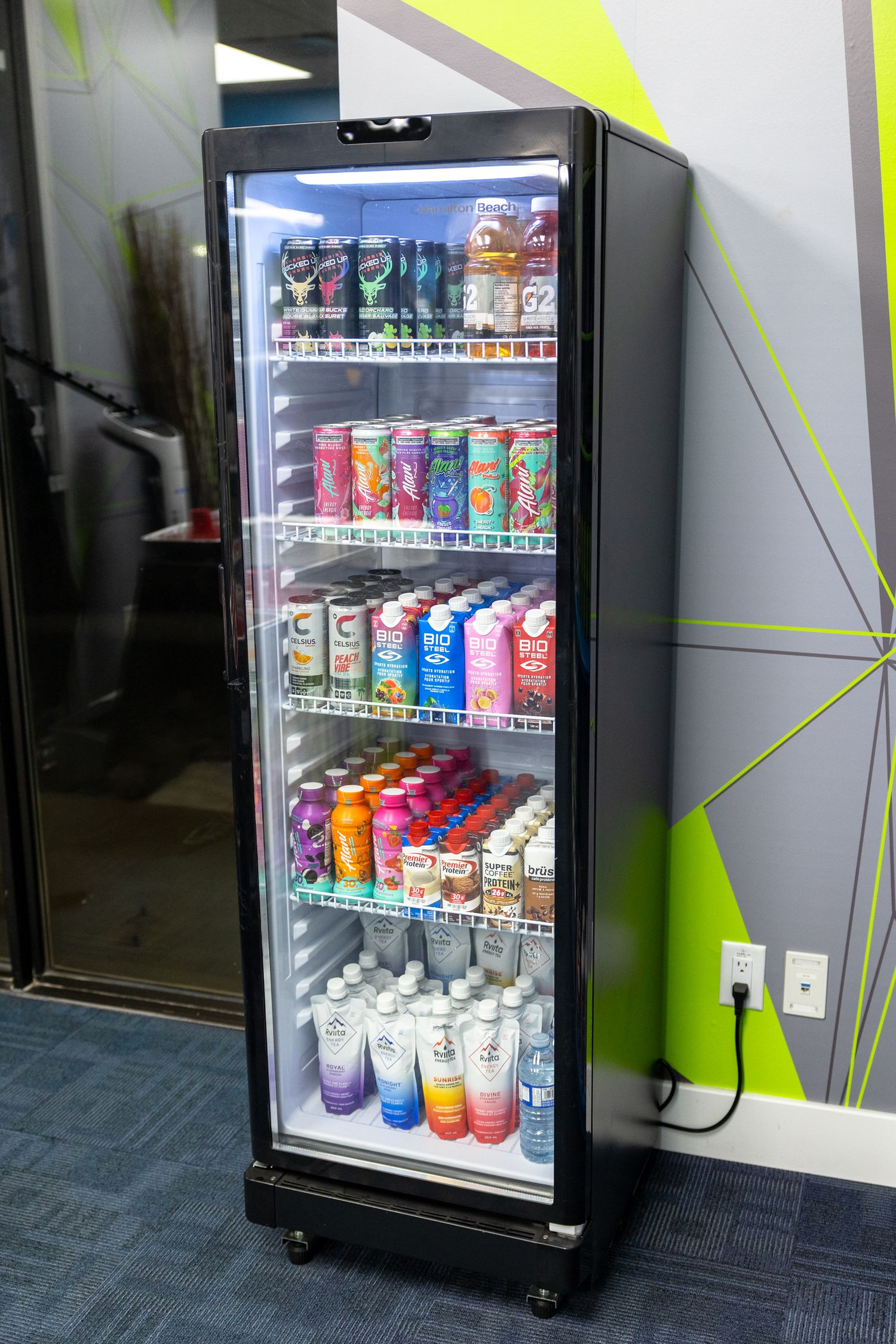A stocked commercial refrigerator filled with colorful canned beverages and bottled drinks on multiple shelves in an office.