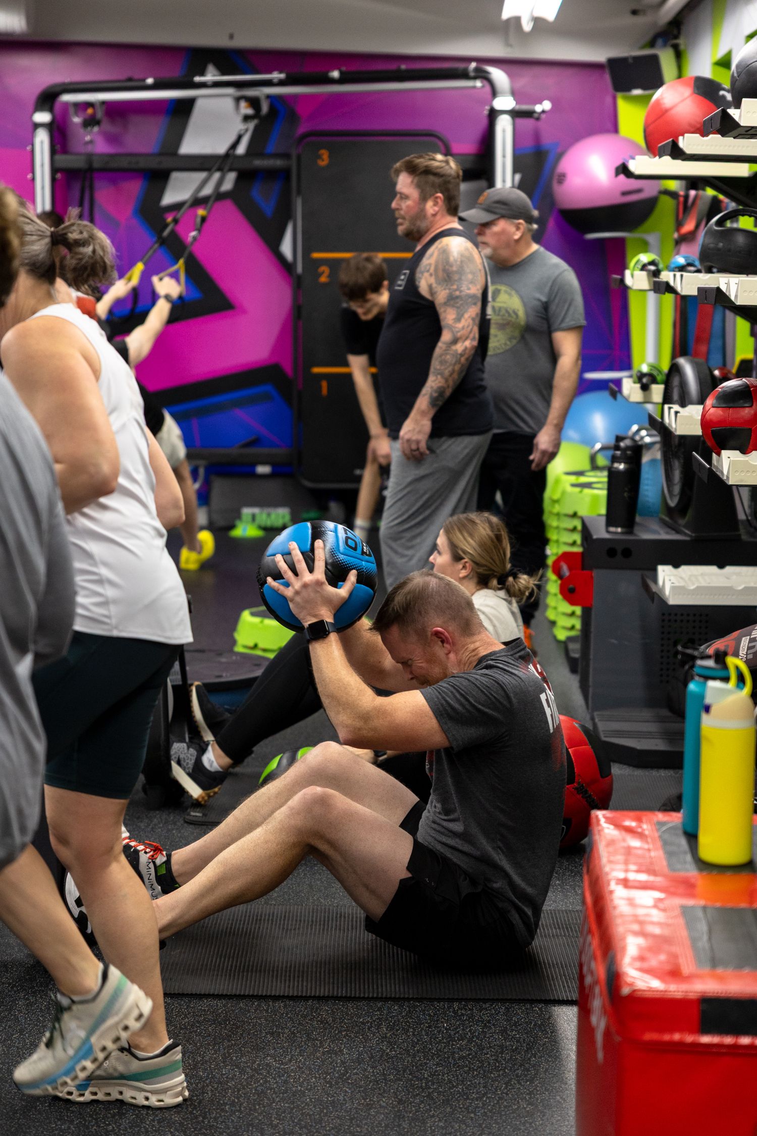 People exercising in a colorful gym, with a man doing sit-ups holding a medicine ball on a red mat.