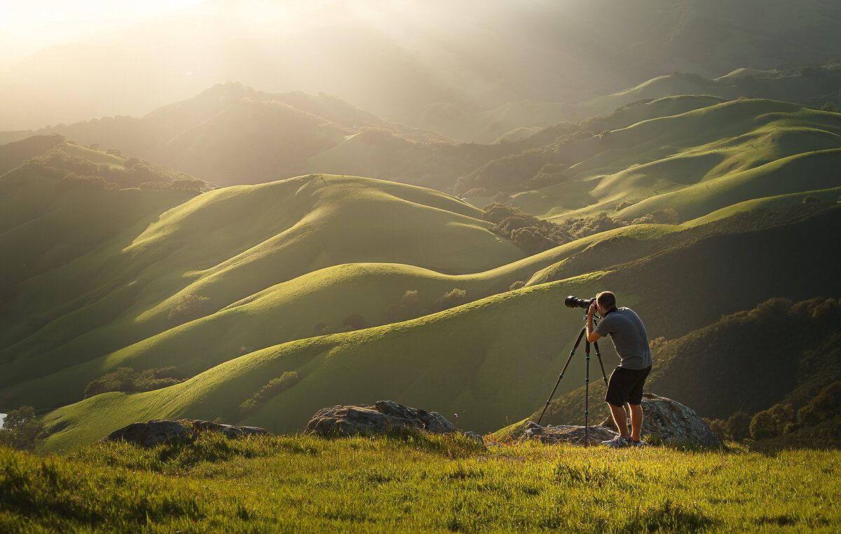 Photographer with tripod captures dramatic sunset over rolling green hills in golden light.