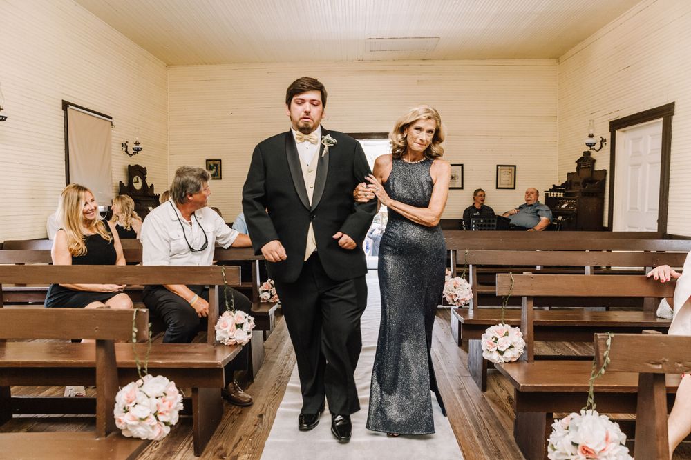 A formal procession through a chapel with wooden pews decorated with white floral arrangements.
