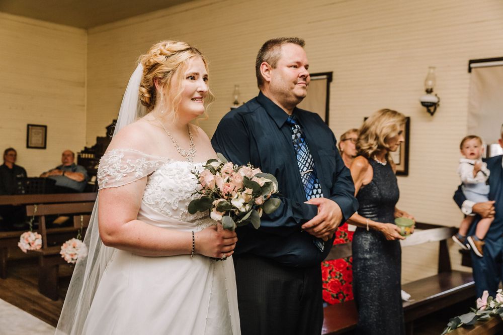 Wedding ceremony processional with couple walking down aisle holding bouquet in rustic chapel.