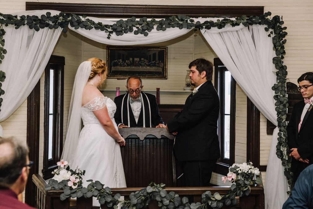 Traditional wedding ceremony under white draped arch with greenery decorations in indoor venue.