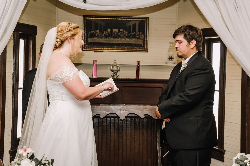 Exchange of vows during wedding ceremony under white draped arch with vintage decor.