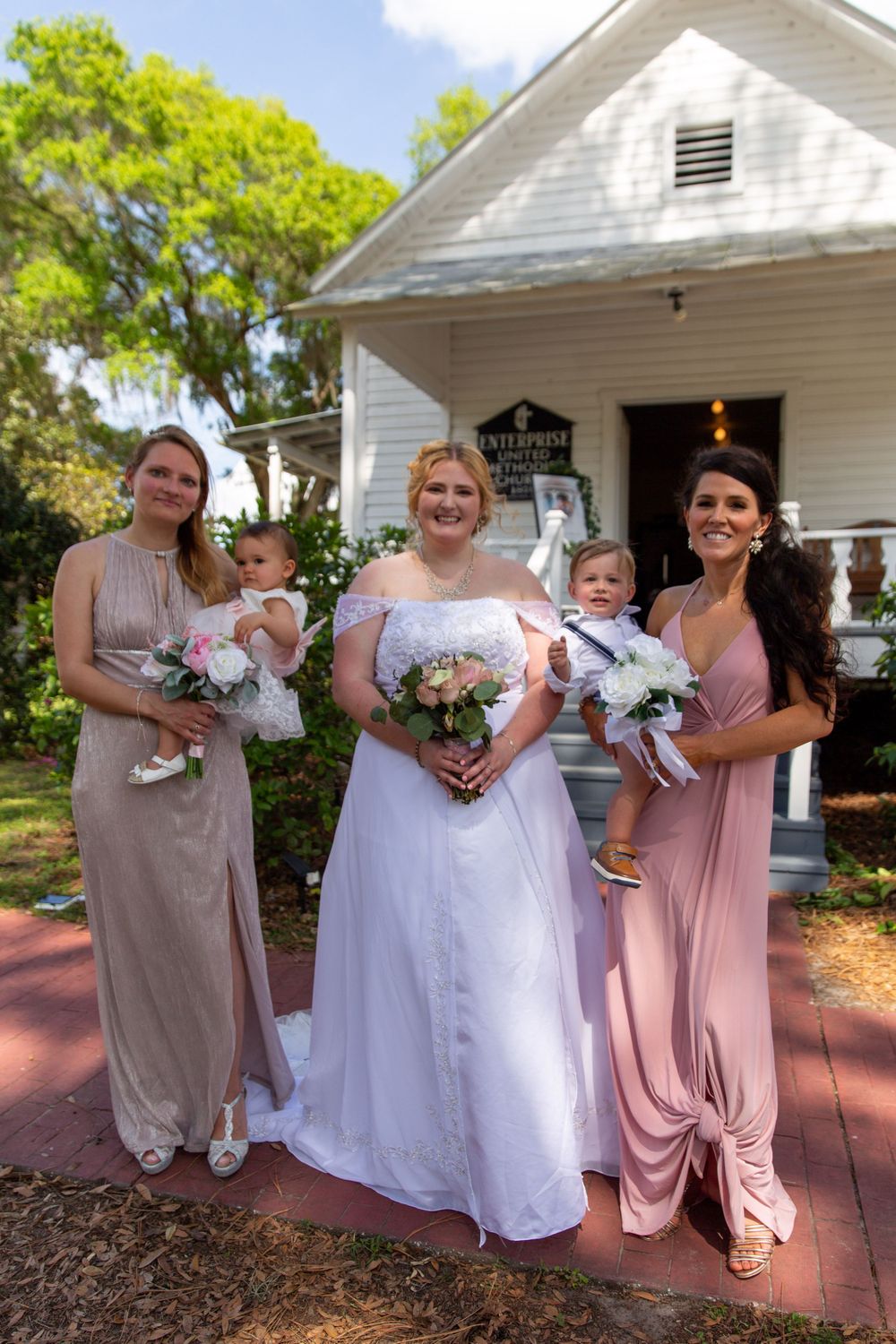 Bridal party poses in front of white chapel with bridesmaids in pink dresses holding infants.