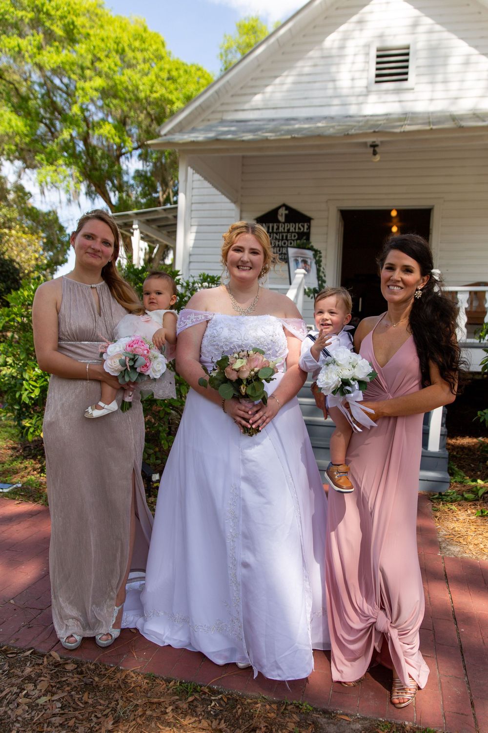Wedding party poses outside historic white chapel with bridesmaids in pink and taupe dresses.