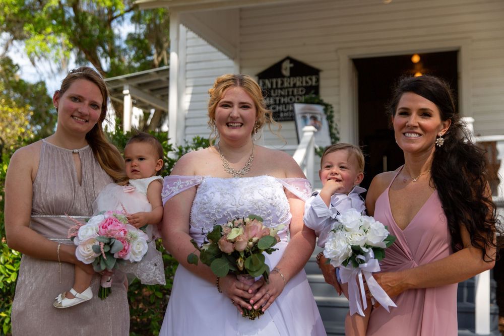 Bridal party portrait with bridesmaids in blush dresses holding babies outside white chapel.
