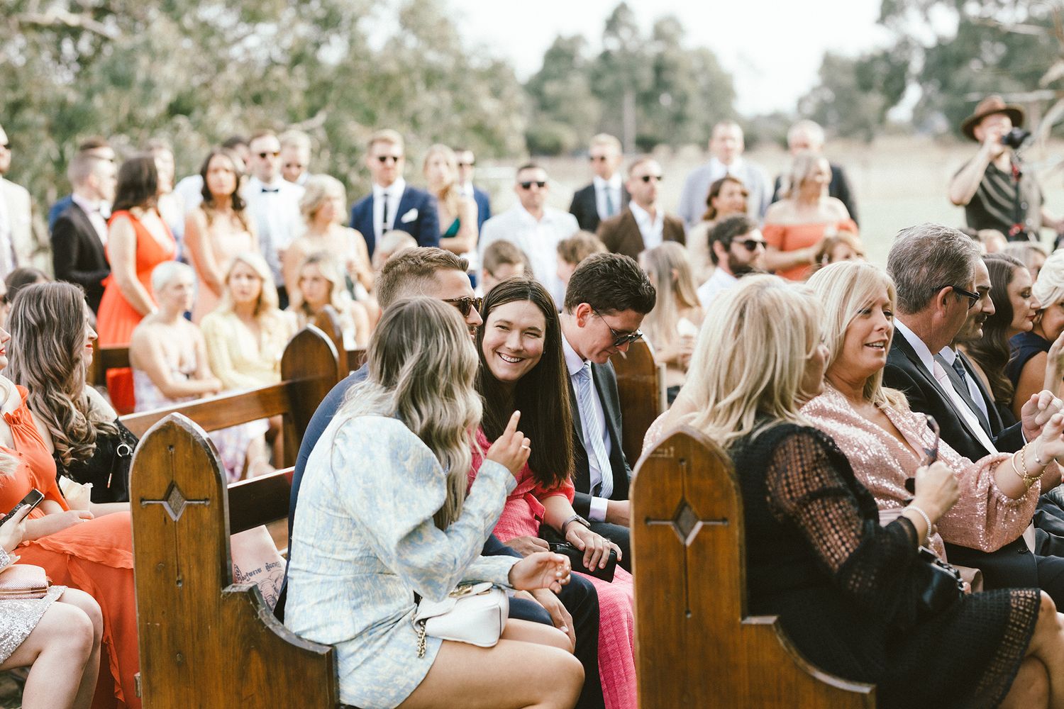 Wedding guests seated on wooden pews during outdoor ceremony.