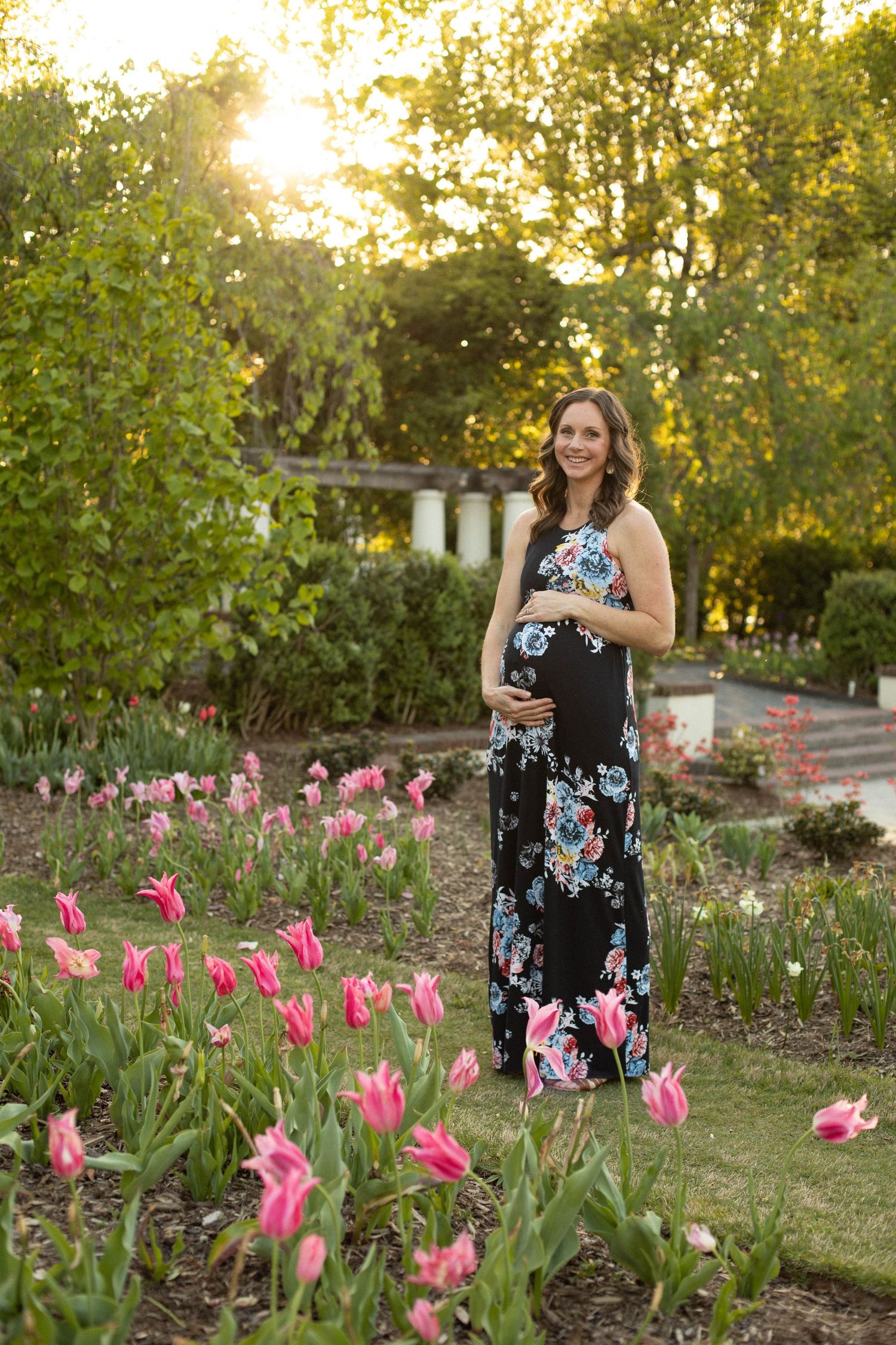 Woman in floral dress poses among pink tulips in a spring garden during golden hour.