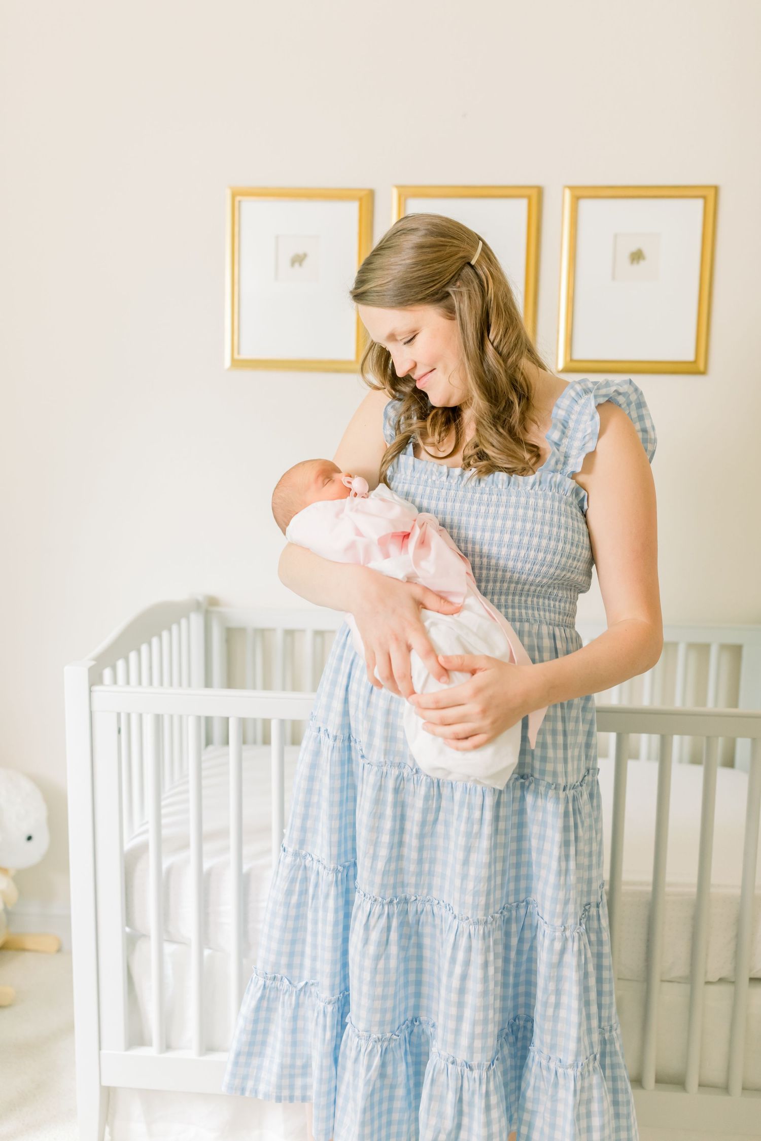 A blue gingham dress-wearing figure tenderly holds a newborn baby in a nursery with gold-framed artwork.