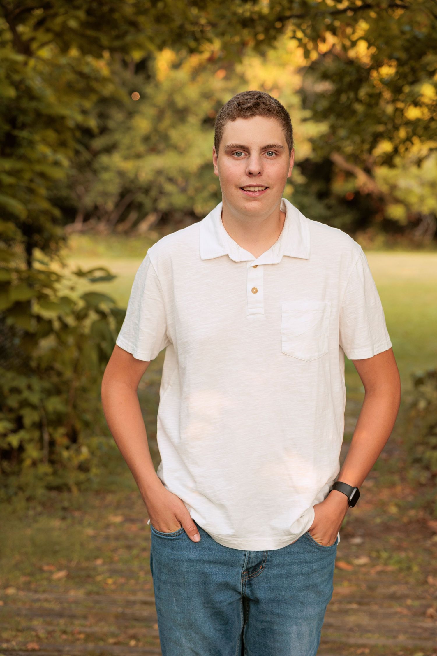 A relaxed outdoor photo of someone in a white polo and jeans with hands in pockets.