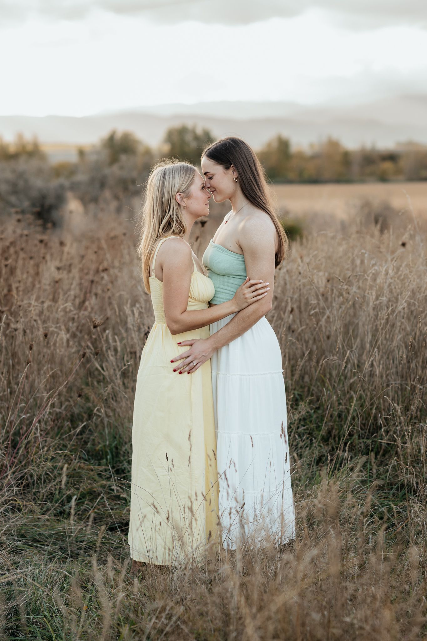 Two women in light colored dresses embrace while standing in a golden sunlit field of tall grass at sunset.