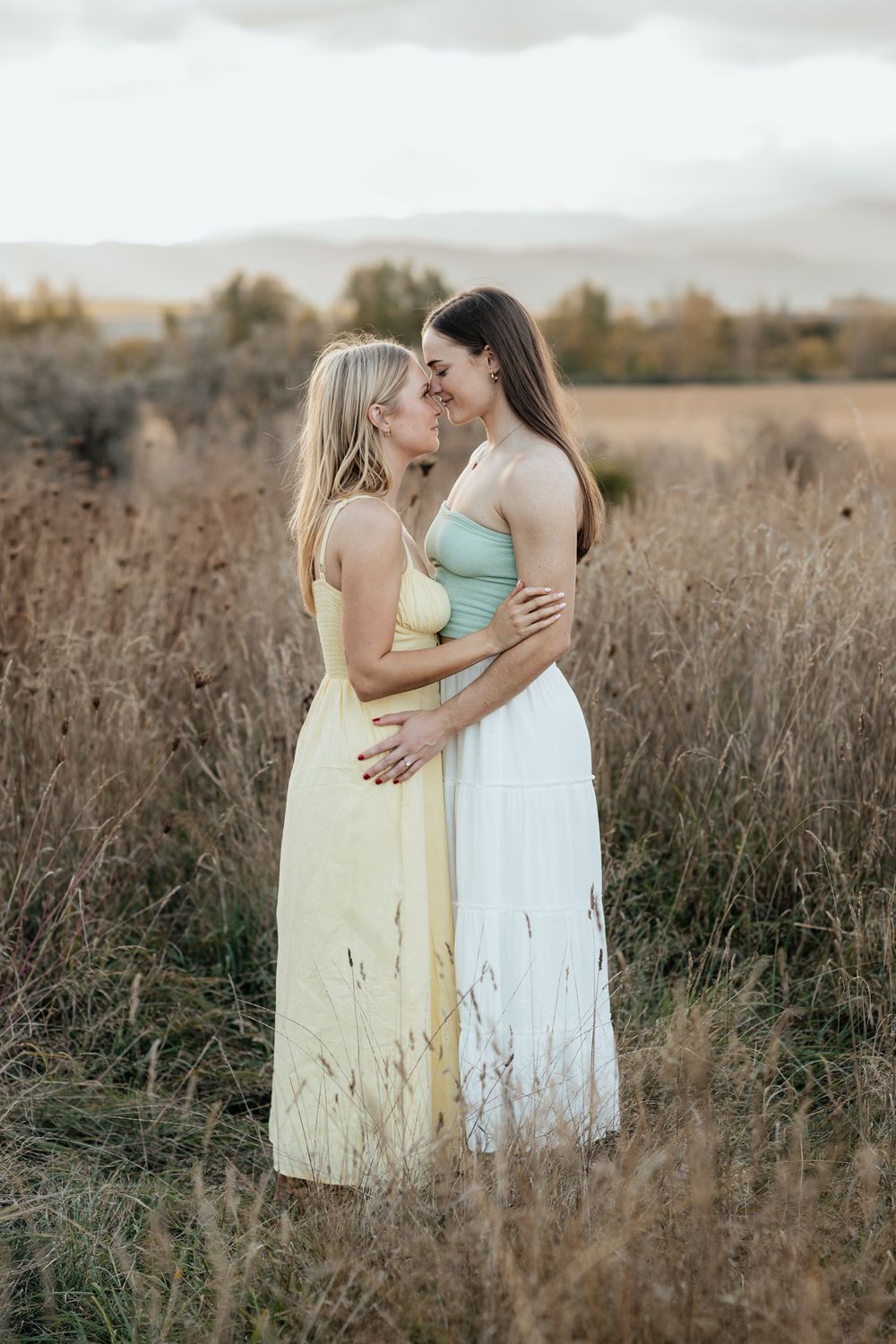 Two women in light colored dresses embrace while standing in a golden sunlit field of tall grass at sunset.