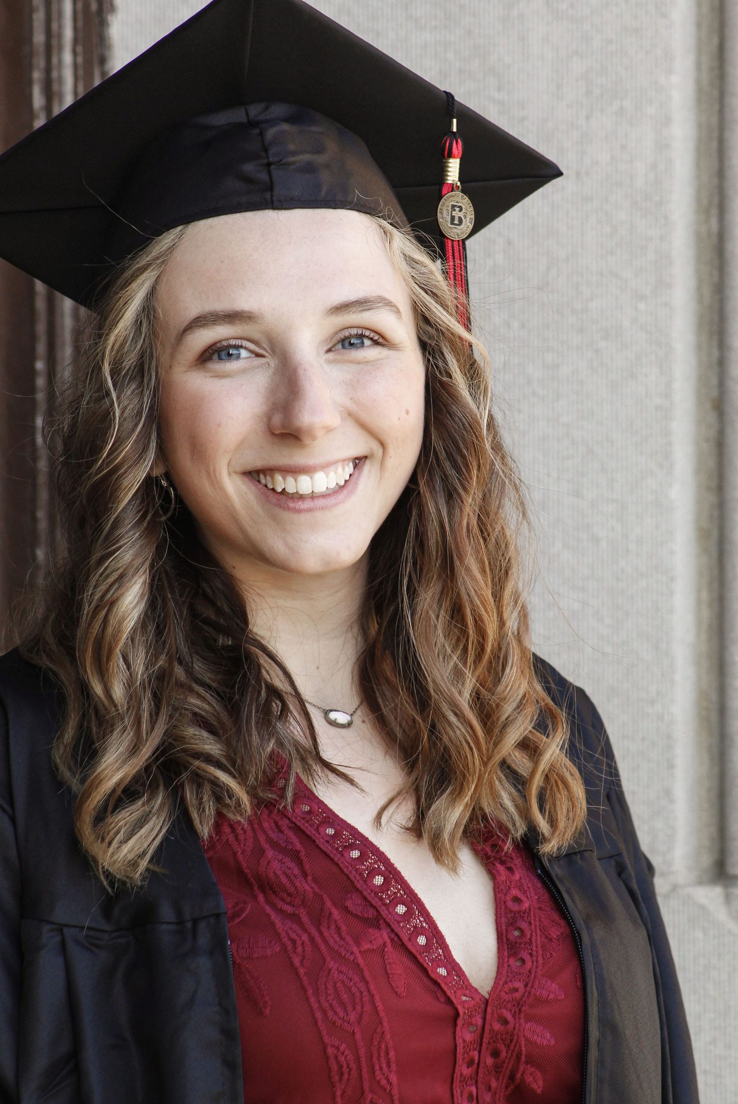 Graduate in burgundy dress and black cap poses for graduation photos.