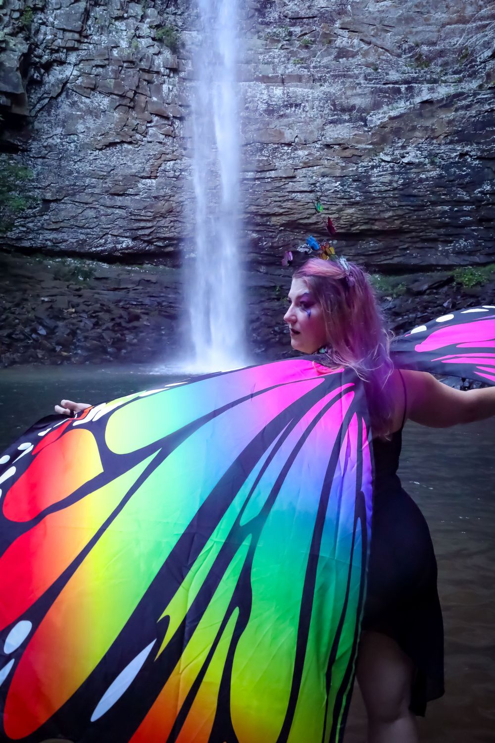 A person wearing rainbow butterfly wings poses dramatically in front of a majestic waterfall.