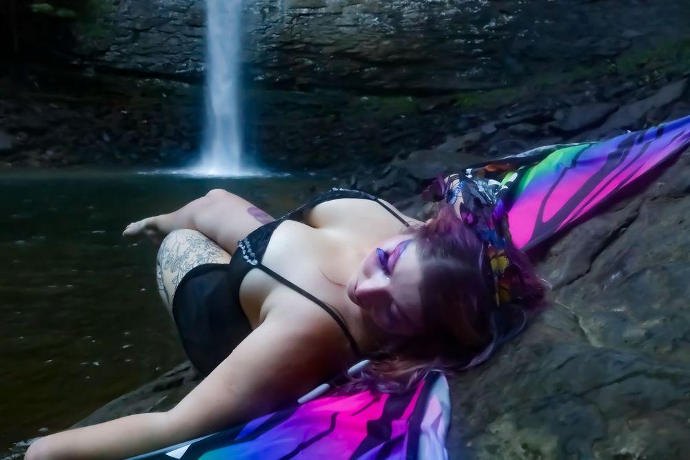 A person with colorful hair lies near a waterfall in artistic boudoir photography.