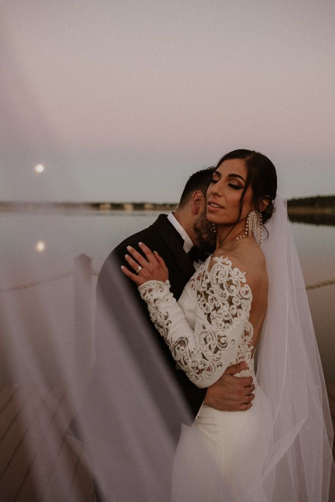 A romantic sunset wedding portrait with a bride in lace sleeves embracing her partner by the water.