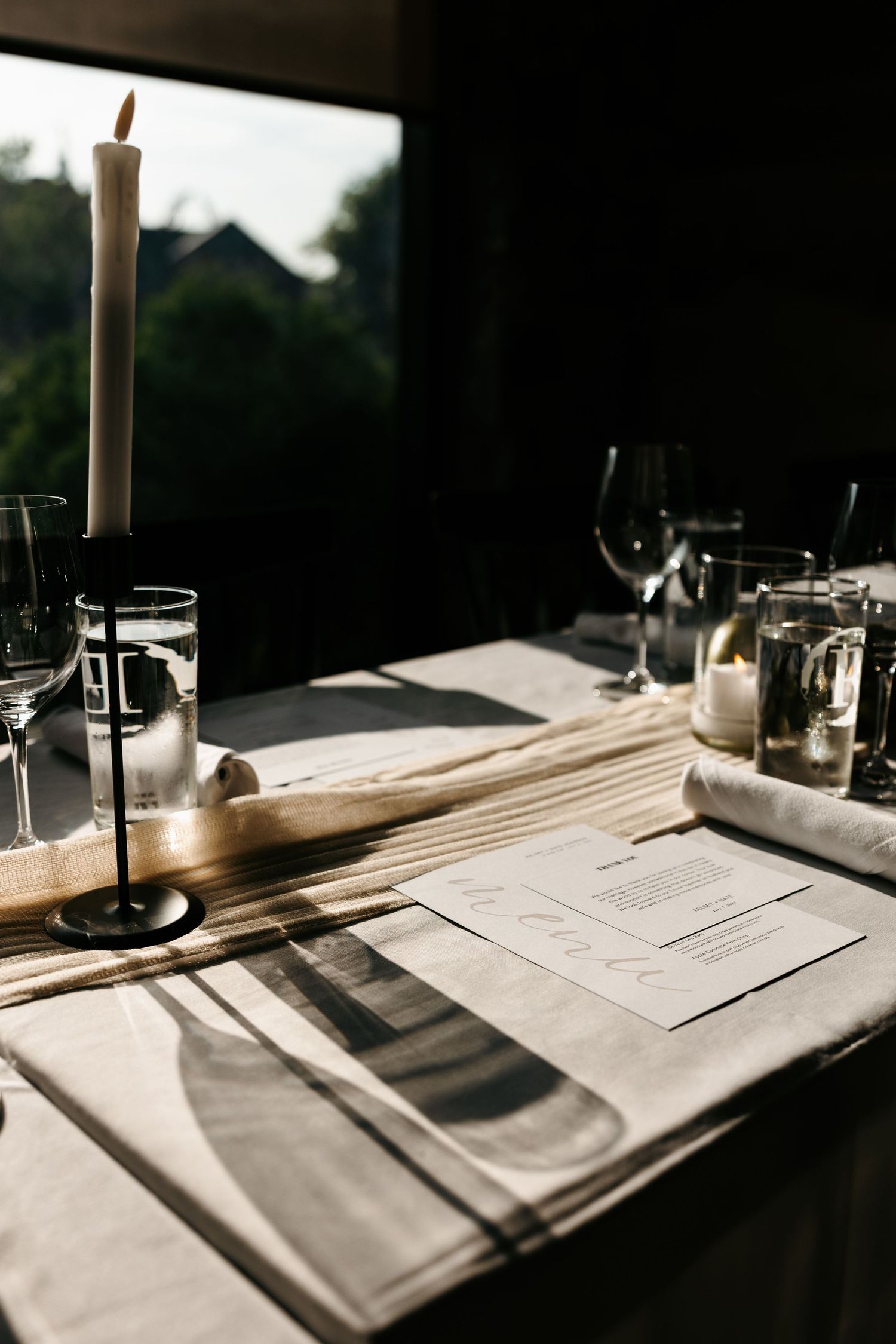 Elegant table setting with white candle, wine glasses and menu cards in moody lighting.