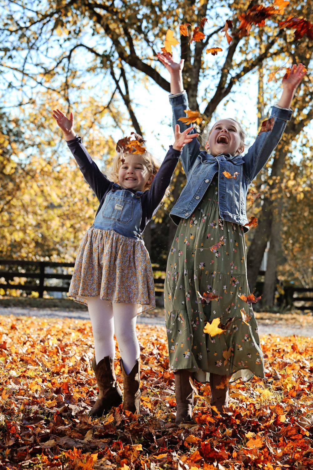Two children in denim jackets throwing autumn leaves in the air while standing on fallen foliage.