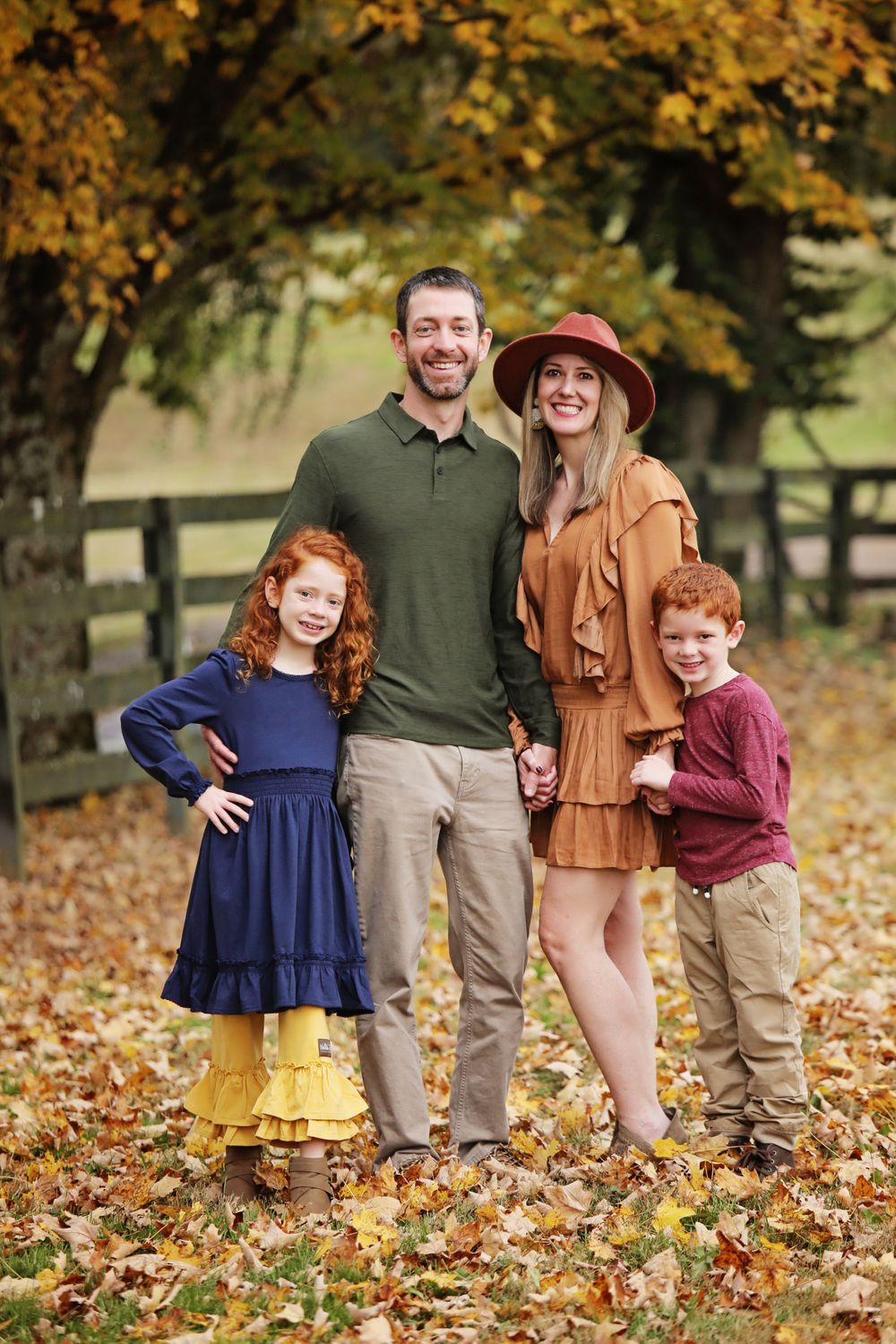 Family of four poses for portrait in autumn setting with rustic fence and golden trees.