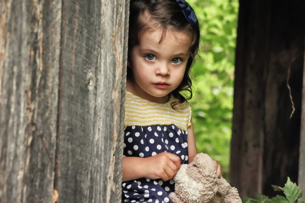 Young child in polka dot dress peeks through wooden structure with stuffed toy in Nolensville photoshoot.