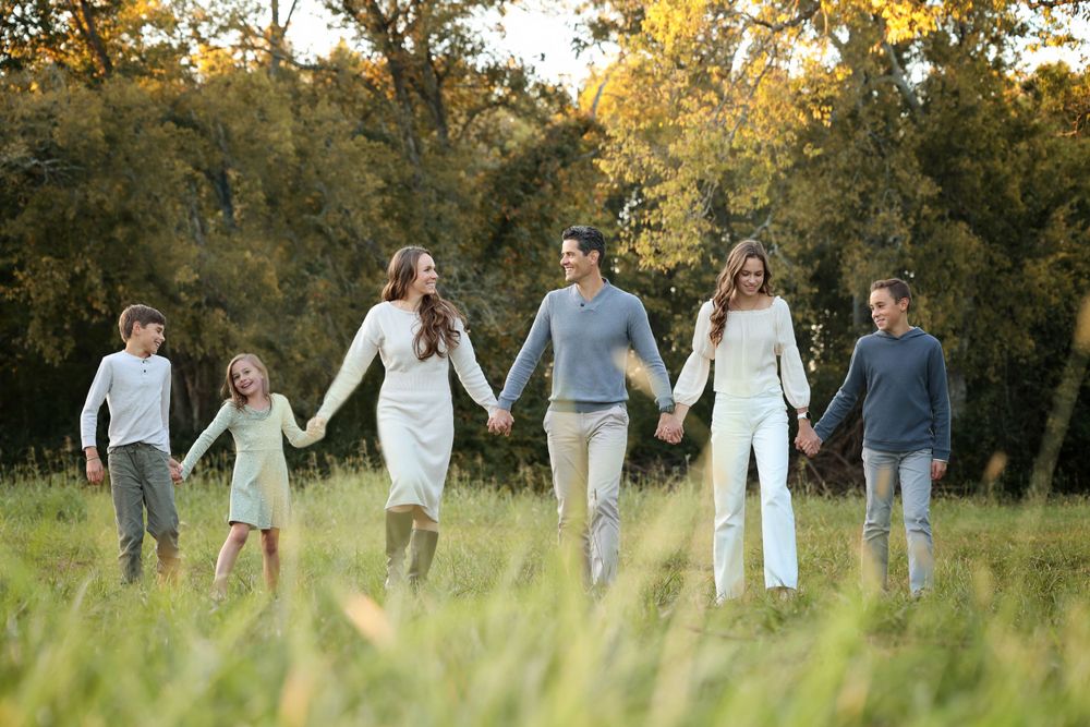 Group walking hand in hand through tall grass field at sunset in coordinated white and gray outfits in custom family photo session outdoors.