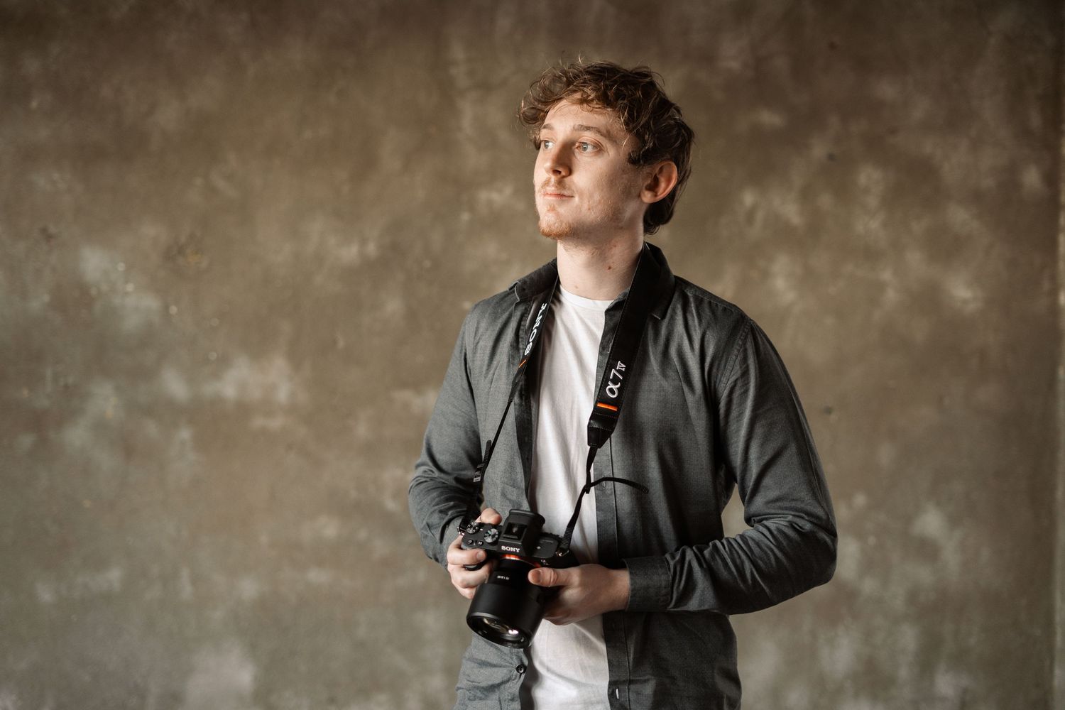 Photographer in gray jacket holds camera while looking up against textured background.